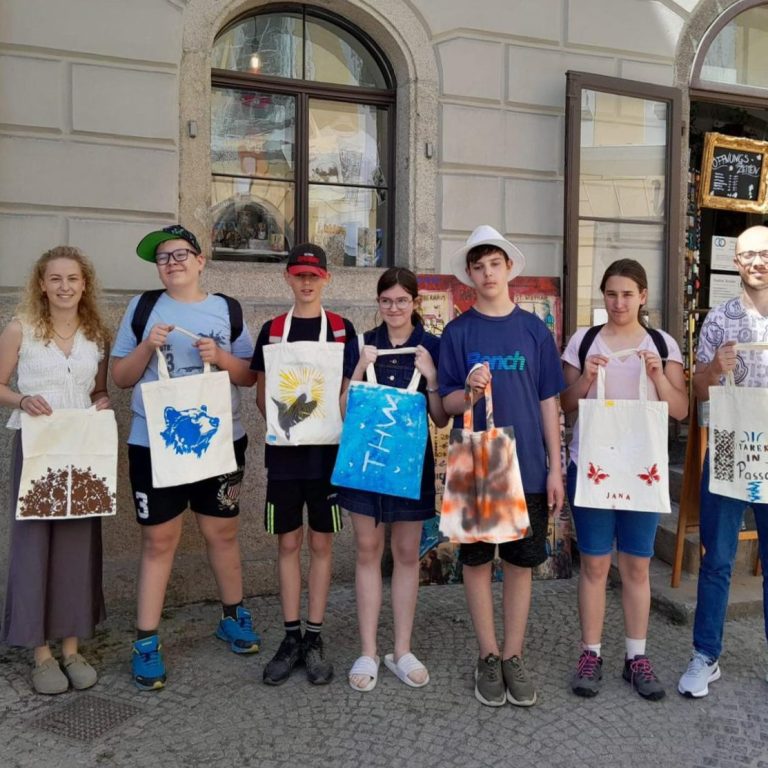 Sechs Personen halten bemalte Stoffbeutel vor einem Geschäft in einer Stadtansicht.