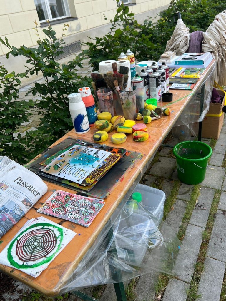 Ein Tisch mit Kunstmaterialien, Farben, Flaschen und Papier in einem Garten.